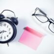 Wecker, Brille und rosa Haftnotiz auf weißem Hintergrund – Symbole für Zeitmanagement und Organisation am Arbeitsplatz.