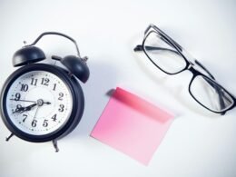 Wecker, Brille und rosa Haftnotiz auf weißem Hintergrund – Symbole für Zeitmanagement und Organisation am Arbeitsplatz.