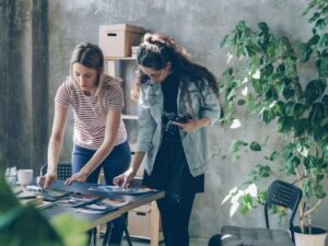 Zwei Frauen sitzen an einem Tisch, diskutieren und arbeiten gemeinsam an Unterlagen in einem modernen Büro