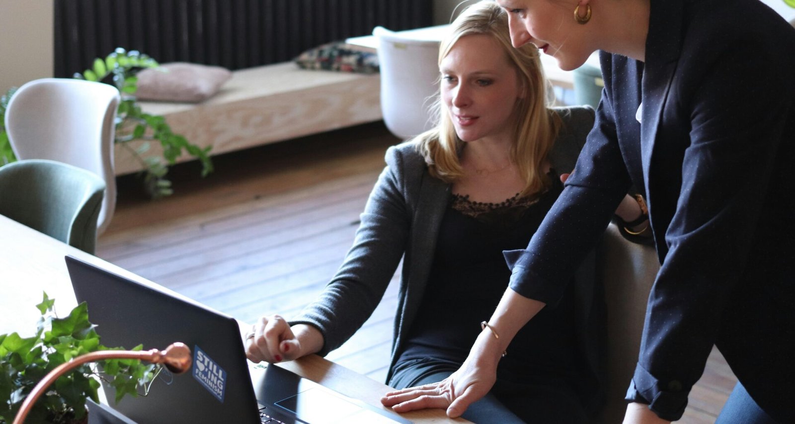 Zwei Frauen sitzen an einem Tisch und schauen gemeinsam auf einen Laptop, Zusammenarbeit im modernen Büroalltag.