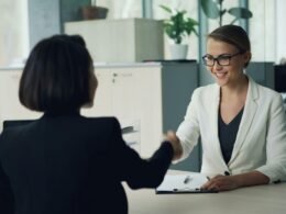 Zwei Frauen sitzen an einem Schreibtisch und schütteln sich zur Begrüßung die Hand in einem modernen Büro