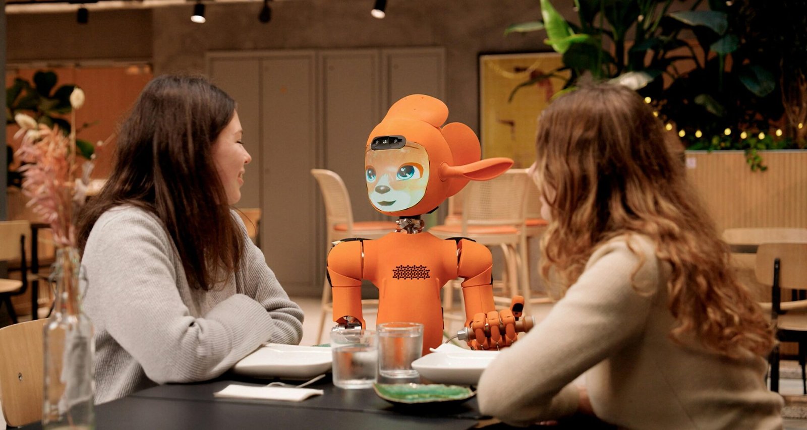 Zwei Frauen sitzen an einem Tisch und sprechen mit einem orangefarbenen Roboter in einer modernen Umgebung