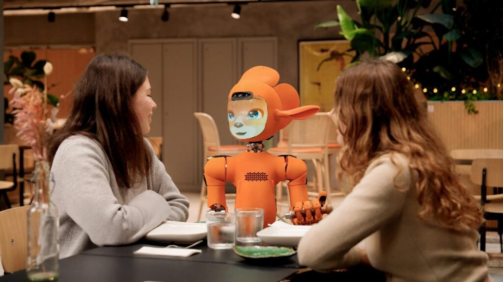 Zwei Frauen führen ein Gespräch mit einem orangefarbenen Roboter an einem Tisch, Fokus auf Technik und Zusammenarbeit
