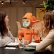 Zwei Frauen sitzen an einem Tisch und unterhalten sich mit einem orangefarbenen Roboter in moderner Umgebung.