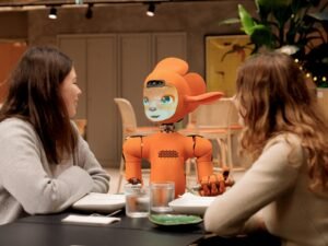 Zwei Frauen sitzen an einem Tisch und unterhalten sich mit einem orangefarbenen Roboter in moderner Umgebung.
