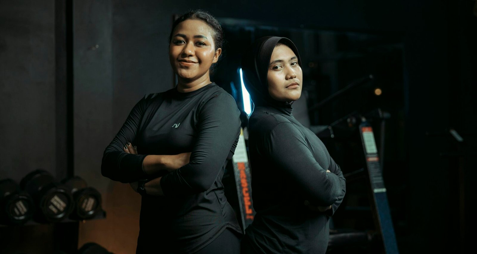 Zwei Frauen stehen in Trainingskleidung nebeneinander in einem modernen Fitnessstudio mit Sportgeräten im Hintergrund.