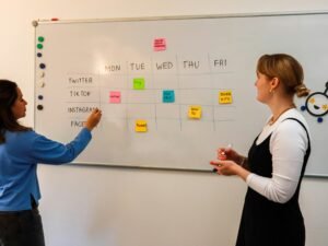 Zwei Frauen diskutieren vor einem Whiteboard mit bunten Klebezetteln in einem modernen Büro.