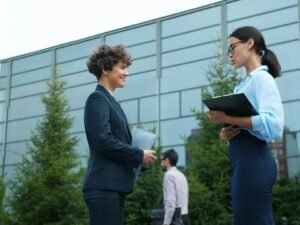 Zwei Geschäftsfrauen unterhalten sich im Freien vor einem modernen Bürogebäude an einem sonnigen Tag.