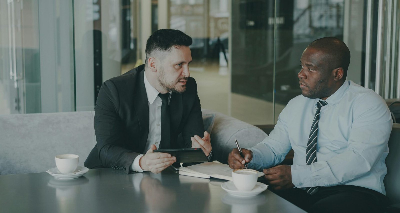 Zwei Männer im Geschäftsgespräch mit Kaffee am Tisch, diskutieren gemeinsam Arbeitsdokumente in modernem Büro