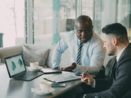 Zwei Geschäftsleute betrachten und diskutieren gemeinsam verschiedene Diagramme auf einem Laptop am Schreibtisch.