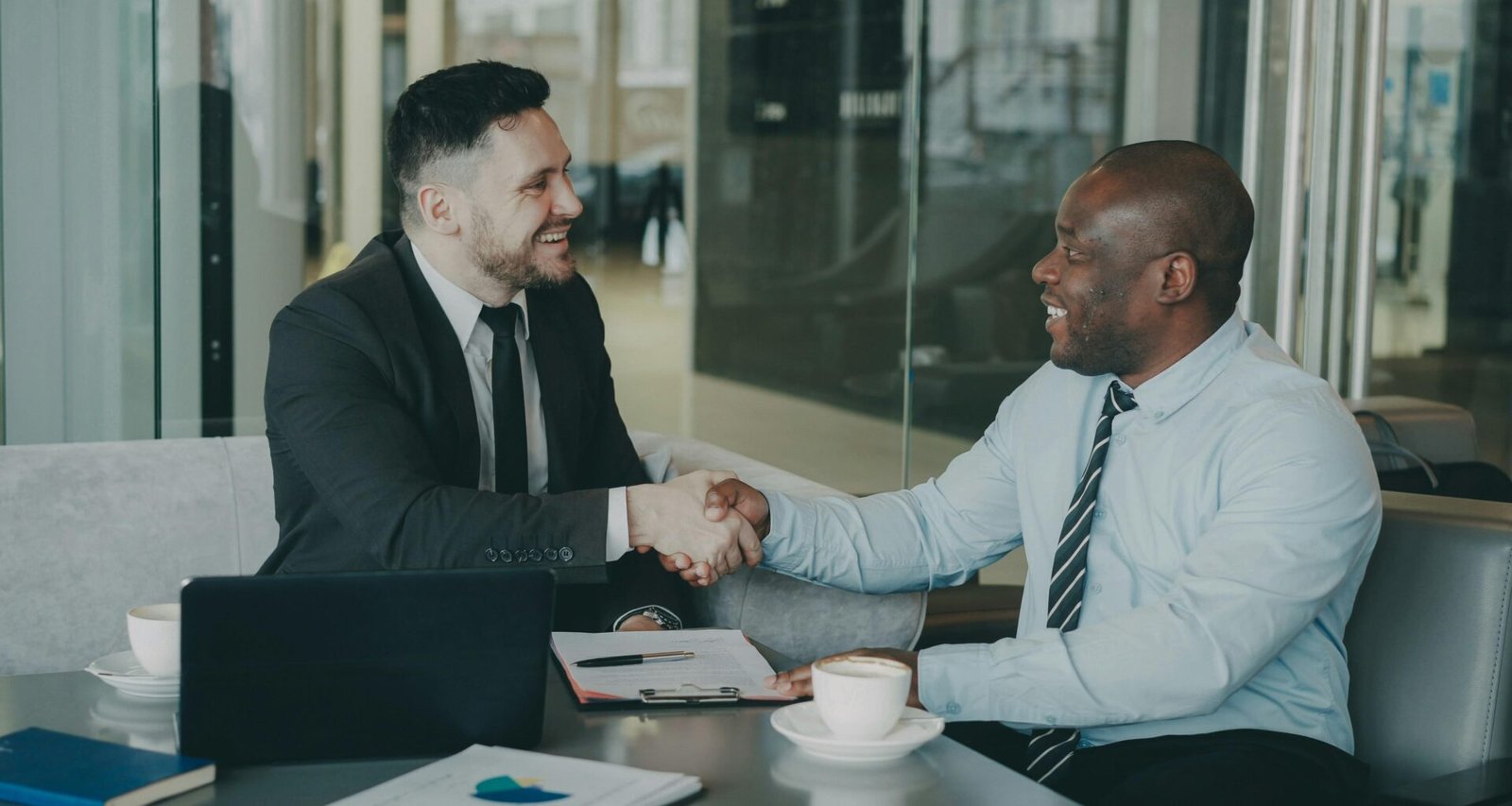 Zwei Männer in Businesskleidung sitzen an einem Tisch und geben sich zur Begrüßung oder zum Abschluss eines Treffens die Hand.