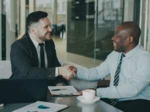Zwei Männer in Businesskleidung sitzen an einem Tisch und geben sich zur Begrüßung oder zum Abschluss eines Treffens die Hand.