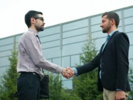 Zwei Geschäftsleute geben sich die Hand vor einem zeitgenössischen Bürogebäude, Symbol für Geschäftspartnerschaften