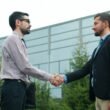 Zwei Männer im Anzug schütteln sich vor einem modernen Bürogebäude die Hand, Symbol für Partnerschaft