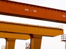 Zwei große Industriekräne stehen nebeneinander vor einem wolkenbehangenen, hellen Himmel.