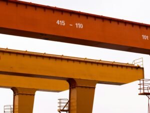 Zwei große Industriekräne stehen nebeneinander vor einem wolkenbehangenen, hellen Himmel.