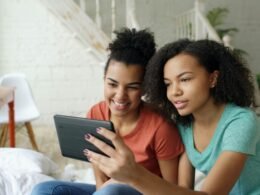 Zwei junge Frauen sitzen nebeneinander und schauen gemeinsam auf ein Tablet, das sie in den Händen halten