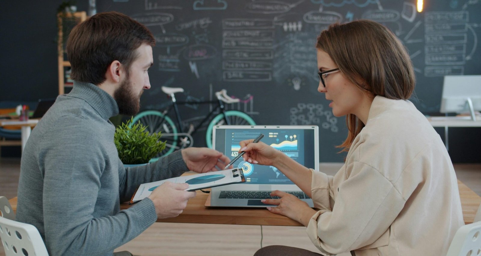 Zwei Kollegen sitzen an einem Tisch und prüfen zusammen Daten auf einem Laptop in einem modernen Büro.