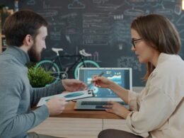 Zwei Kollegen sitzen an einem Tisch und prüfen zusammen Daten auf einem Laptop in einem modernen Büro.