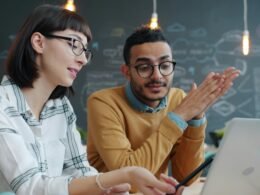 Zwei Kollegen arbeiten gemeinsam an einem Laptop im Büro und tauschen Ideen für ein Projekt aus