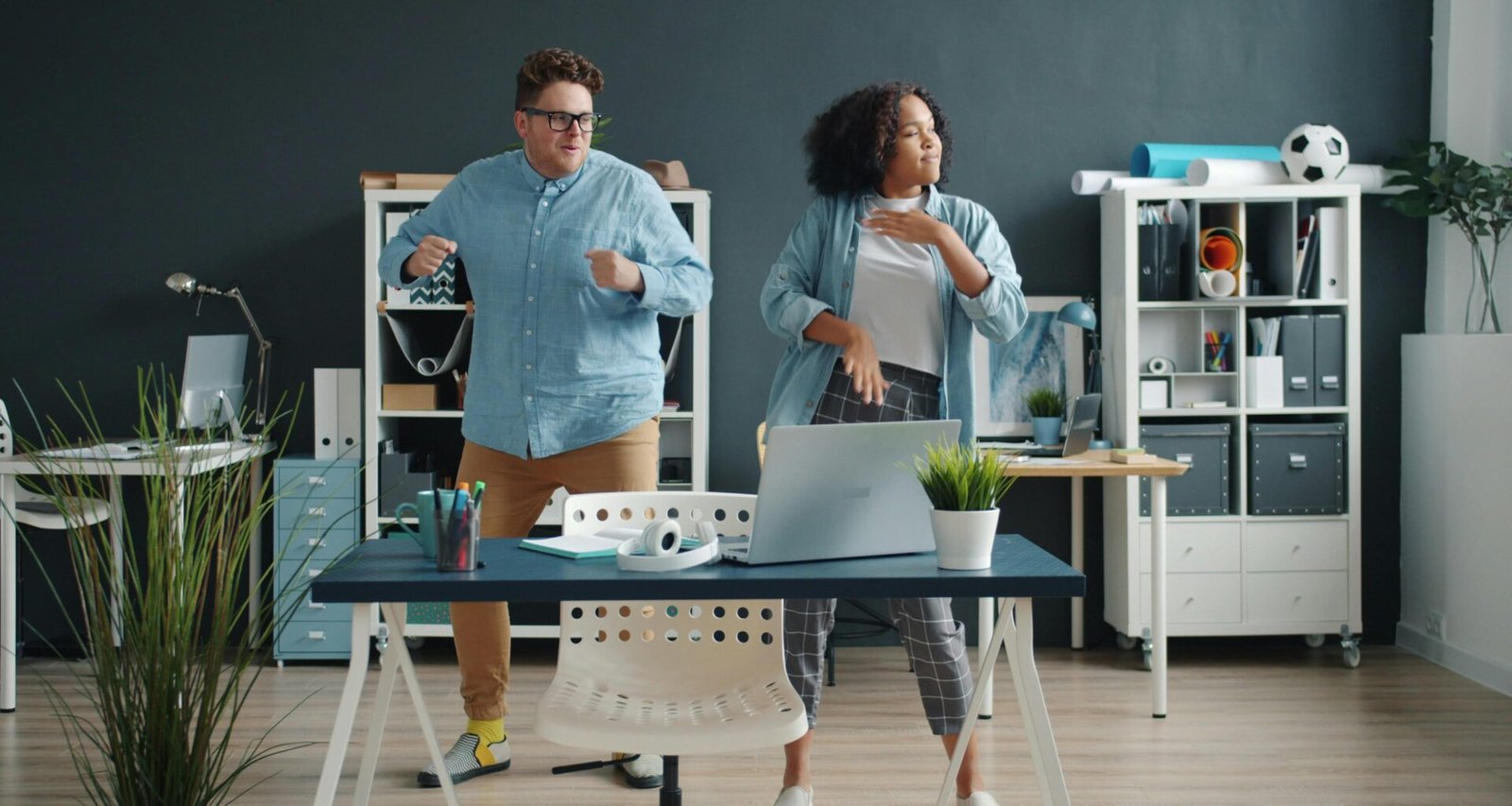 Zwei Kollegen tanzen ausgelassen in einem modernen Büro mit großen Fenstern und Schreibtischen im Hintergrund.