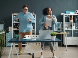 Zwei Kollegen tanzen ausgelassen in einem modernen Büro mit großen Fenstern und Schreibtischen im Hintergrund.
