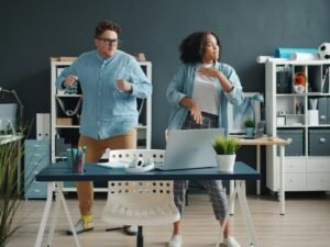 Zwei Kollegen tanzen ausgelassen in einem modernen Büro mit großen Fenstern und Schreibtischen im Hintergrund.