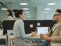 Zwei Personen sitzen an Schreibtischen und diskutieren gemeinsam Arbeitsaufgaben im modernen Büro