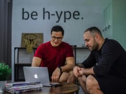 Zwei Männer sitzen an einem Kaffeetisch und schauen gemeinsam auf einen Laptop, konzentriert im Gespräch über digitale Themen.