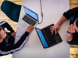 Zwei Männer sitzen sich an einem Tisch gegenüber und arbeiten konzentriert an ihren Laptops in einem modernen Büro.