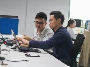 Zwei Männer sitzen an einem Schreibtisch und betrachten gemeinsam den Bildschirm eines Laptops in einem modernen Büro.