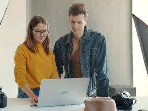 Zwei Menschen sitzen nebeneinander in einem Studio und schauen gemeinsam auf den Bildschirm eines Laptops.
