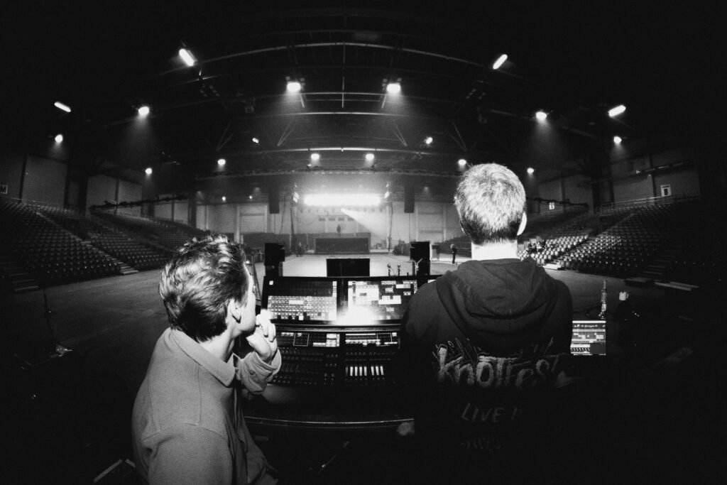 Zwei Personen sitzen in einer leeren Arena und schauen auf eine beleuchtete Bühne, die sich vorne befindet.