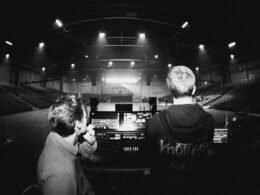 Zwei Personen sitzen in einer leeren Arena und schauen auf eine beleuchtete Bühne, die sich vorne befindet.