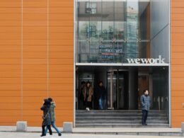 Zwei Personen spazieren auf dem Gehweg vor einem modernen Wework-Gebäude in einer Stadtumgebung