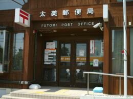 Detaillierte Aufnahme eines Futomi Postamt-Schildes mit japanischen Schriftzeichen und Symbol vor einfarbigem Hintergrund