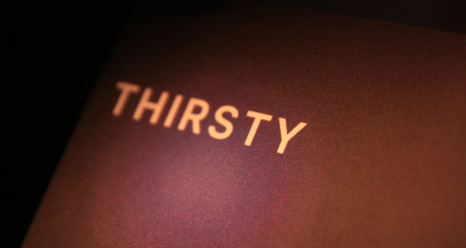 Das Wort "thirsty" steht weiß geschrieben auf einer dunkel gehaltenen Oberfläche, minimalistisch und klar fotografiert.