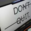 Ein Computerbildschirm zeigt den Text 'don't quit.' auf dunklem Hintergrund, Schreibtisch im modernen Büro