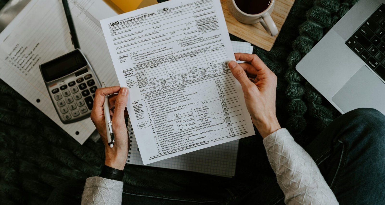 Hände halten Steuerformulare neben Taschenrechner und Laptop auf einem Schreibtisch, Fokus auf digitale Bearbeitung