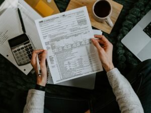 Hände halten Steuerformulare neben Taschenrechner und Laptop auf einem Schreibtisch, Fokus auf digitale Bearbeitung