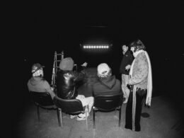 Mehrere Personen sitzen in einem dunklen Raum und schauen aufmerksam auf einen großen beleuchteten Bildschirm.