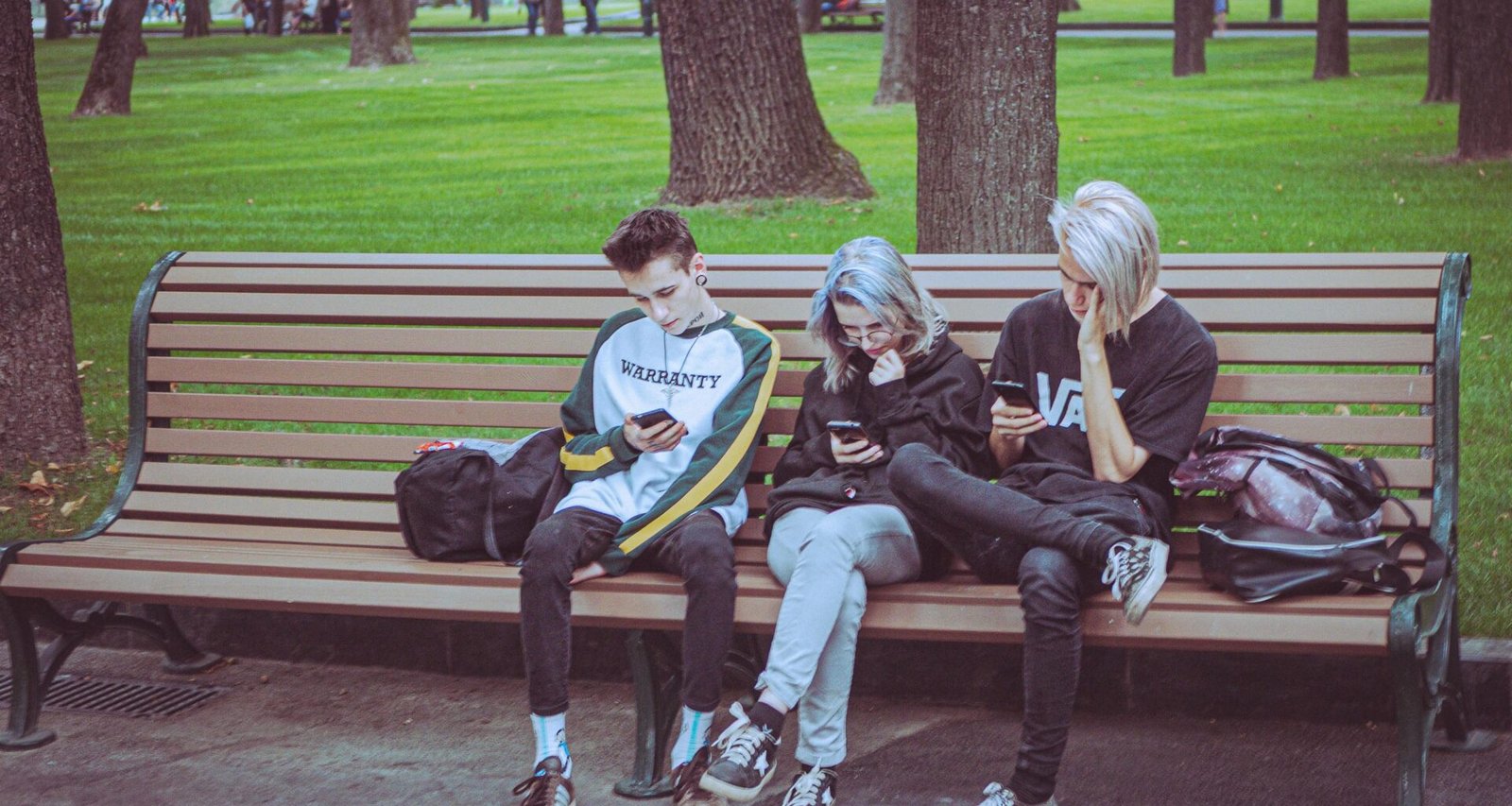 Drei junge Menschen sitzen nebeneinander auf einer Parkbank im Freien und schauen auf ihre Smartphones.