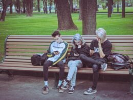Drei junge Menschen sitzen nebeneinander auf einer Parkbank im Freien und schauen auf ihre Smartphones.