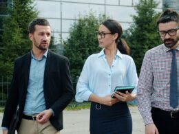 Drei Personen in Businesskleidung laufen gemeinsam vor einem zeitgemäßen Bürogebäude entlang dem Gehweg