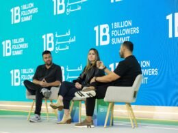 Drei Menschen sitzen auf Stühlen und nehmen an einer Diskussionsrunde auf einer Bühne teil.