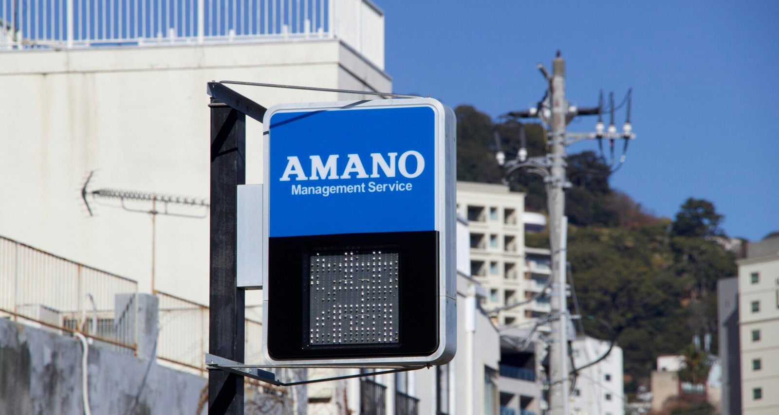Blaues Schild mit Amano Management Service Logo, fotografiert draußen an einer Gebäudefassade bei Tageslicht
