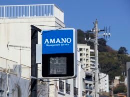 Blaues Schild mit Amano Management Service Logo, fotografiert draußen an einer Gebäudefassade bei Tageslicht