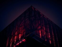 Ein modernes, hohes Gebäude mit rotem Lichtband an der Seitenfassade vor neutralem Himmel.