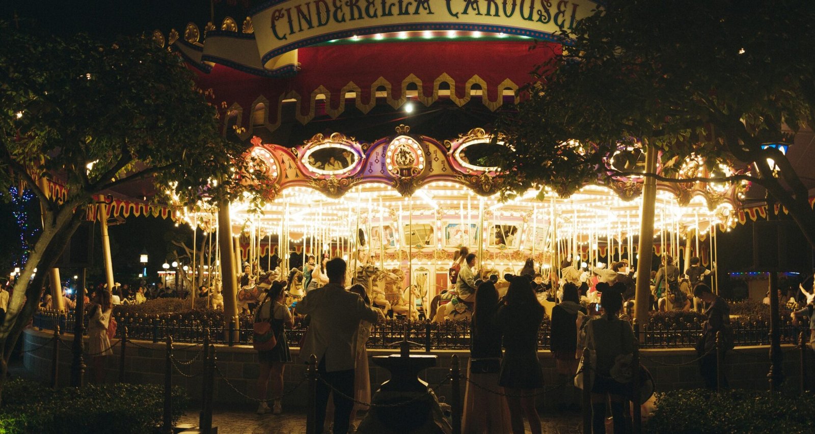 Ein beleuchtetes Karussell bei Nacht, umringt von stehenden Menschen auf einem öffentlichen Platz