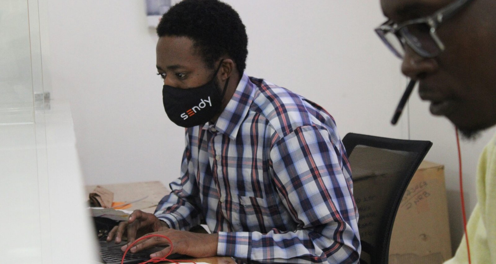 Ein Mann sitzt mit medizinischer Maske am Schreibtisch und arbeitet an einem Laptop in einem modernen Büro.